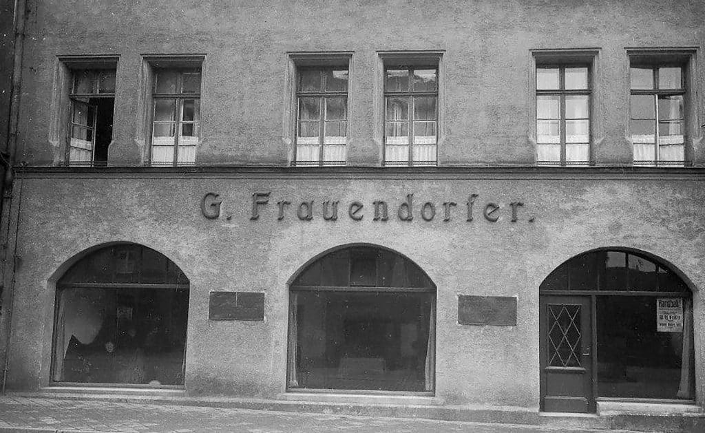 Eine Gebäudefassade mit dem Zeichen G. Frauendorfer über zwei großen Schaufenstern und einer Tür.
