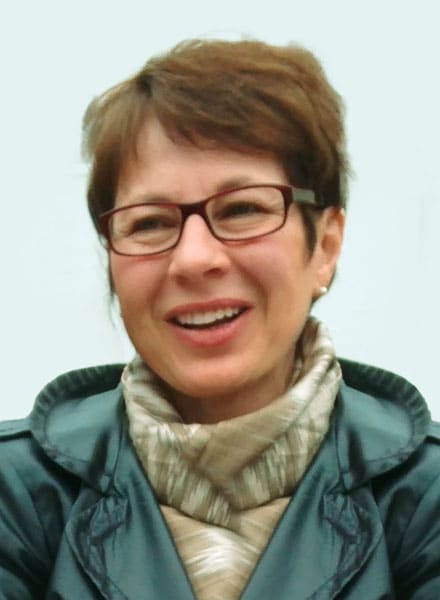 Frau mit kurzen braunen Haaren, Brille und Schal, lächelnd und in einer dunkelgrünen Jacke vor hellem Hintergrund.
