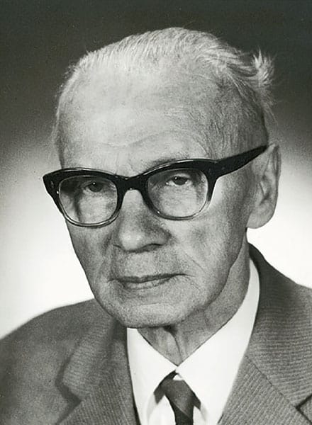 Älterer Mann mit Brille, Anzug und Krawatte, der in einem Schwarz-Weiß-Porträt leicht zur Seite schaut.