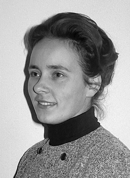 Schwarz-Weiß-Porträt einer Frau mit dunklem Haar, die einen Rollkragenpullover und eine strukturierte Jacke trägt und zur Seite schaut.