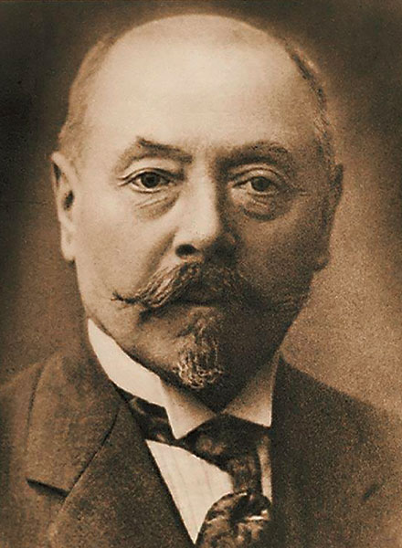 Sepiafarbenes Porträt eines älteren Mannes mit Schnurrbart und Spitzbart, der Anzug und Krawatte trägt und in die Kamera schaut.