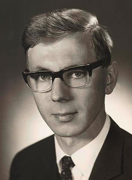 Schwarz-Weiß-Porträt eines jungen Mannes mit Brille, Anzug und Krawatte, der in die Kamera schaut.