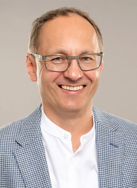 Lächelnder Mann mit Brille, der ein weißes Hemd und einen gemusterten Blazer vor einem schlichten hellen Hintergrund trägt.