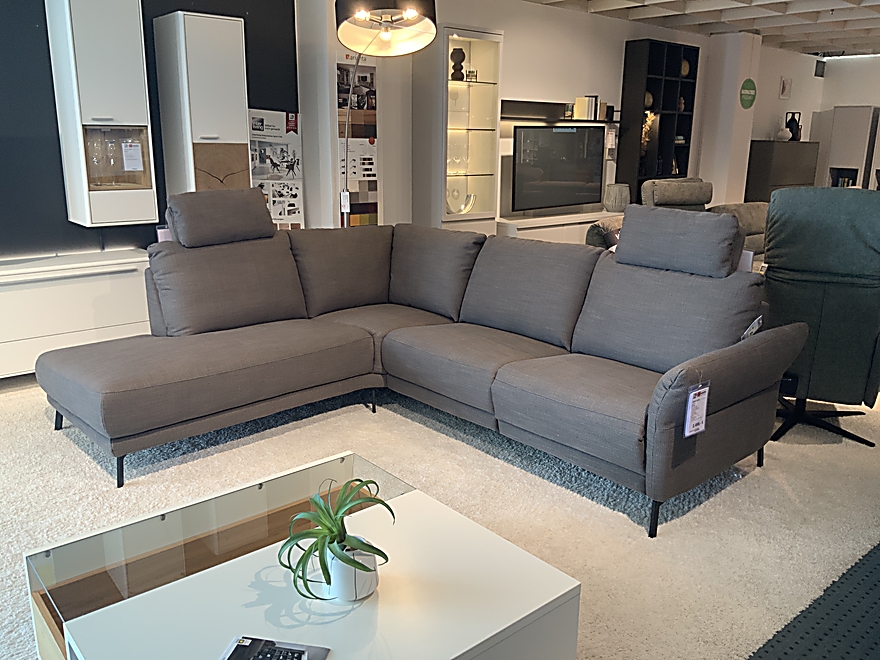 Garnitur Demian grau L-förmigen Sektional in modernen Showroom neben weißen Couchtisch mit Pflanze und Fernbedienung.